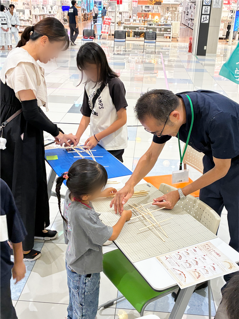 スタッフの説明を受けながら、割り箸を組み合わせてダヴィンチの橋づくりに挑戦する来場者