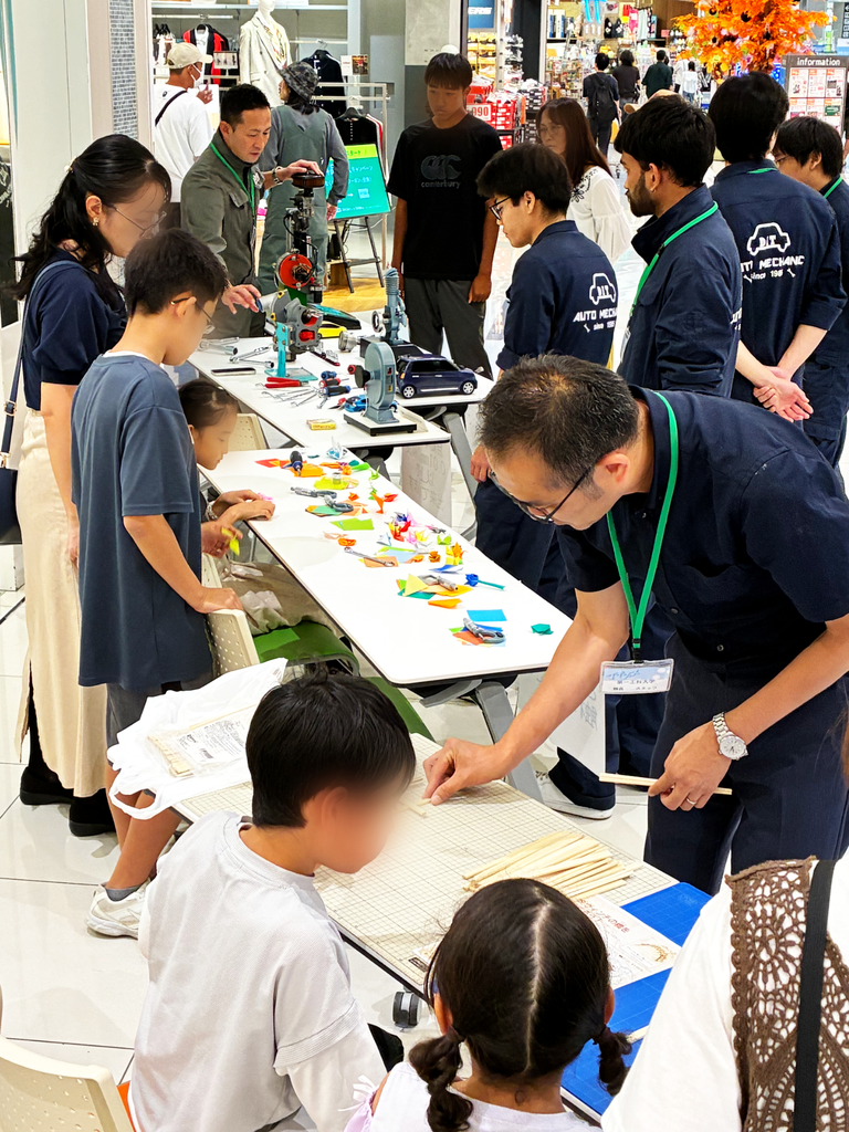 手前で割り箸の橋づくりを体験し、奥で自動車の模型やエンジンの展示を見学する来場者