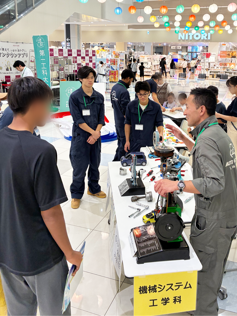 機械システム工学科のブースで、エンジンカットモデルや自動車整備工具を来場者に説明する様子