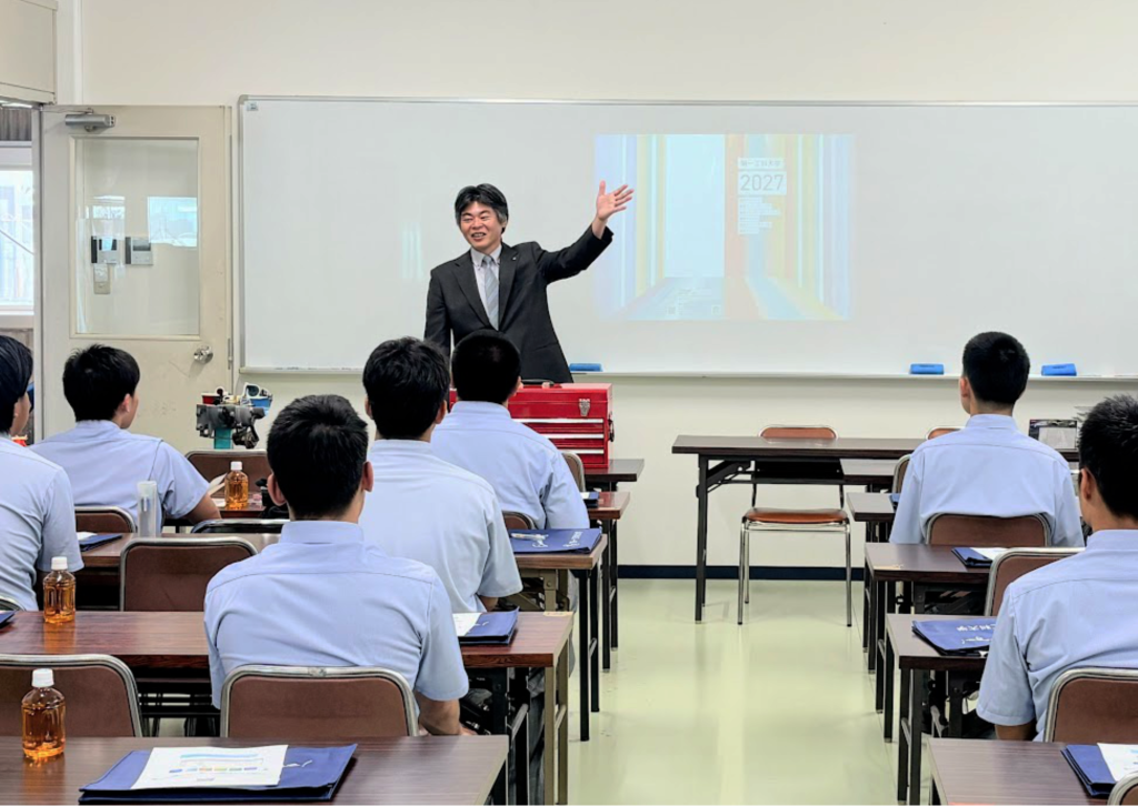 自動車実習棟で歓迎の挨拶をする山田副学長と、着席して話を聞く生徒たち